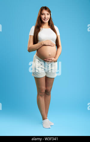 Beautiful pregnant woman in white T-shirt, shorts touching her belly. isolated over a blue background. lifestyle, health care Stock Photo