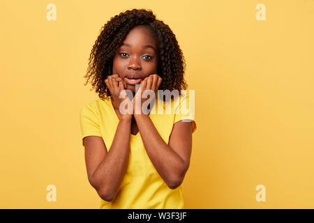 scared emotional american woman keeps , hands on cheeks, being shocked to listen to new, stares at camera, isolated over yellow background. girl in ca Stock Photo