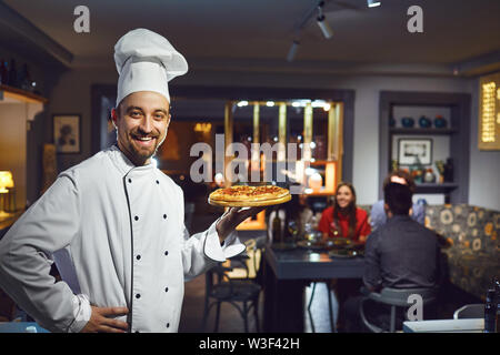 A male cook is smiling against the background of people at a table in a restaurant. Stock Photo