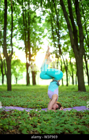 Thin brunette girl  standing in headstand exercise, Garuda salamba sirsasana pose  in a summer park on a sunset background. Woman doing exercises on t Stock Photo