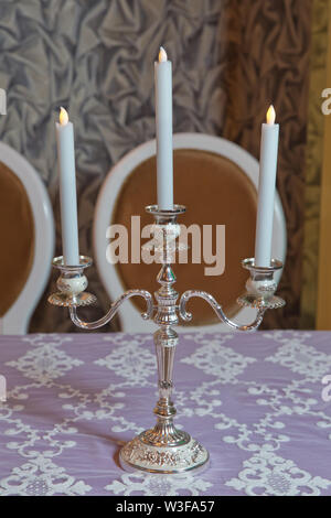 Three white candles on the table . three arms candle stick holder, two candelabra atop, on top of furniture, table against white wall in living room, Stock Photo