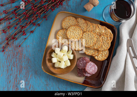 view from above of a glass of red wine with salami and crackers on a plate and cubes of cheese in a little wooden bowl on a teal blue rustic wood back Stock Photo