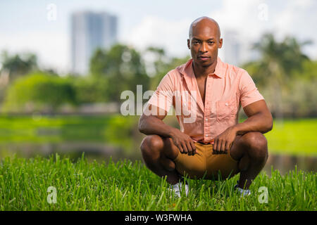 Photo of a handsome young African American man squatting on grass in the park. Deadpan expression staring deep into the camera Stock Photo