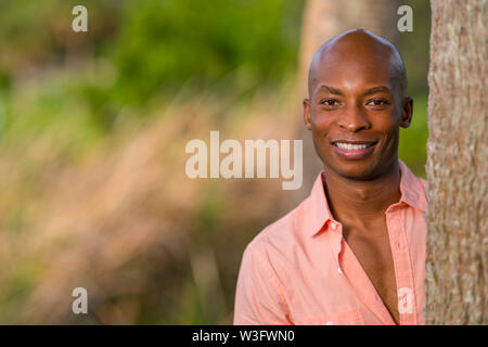 Handsome young African American man posing by a tree in the park. Man wearing a pink button shirt and smiling at the camera Stock Photo