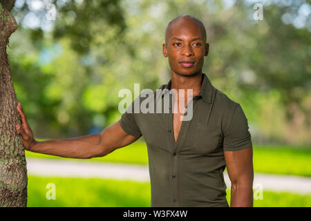 Handsome young African American man posing with hand on tree in the park. Mans shirt is unbuttoned to show chest as man glances away from camera Stock Photo