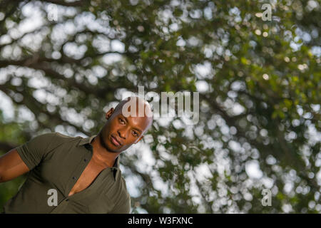 Offset photo of a handsome young black man posing with green trees blurry bokeh background. Man is posing in a retro vintage shirt unbuttoned with big Stock Photo
