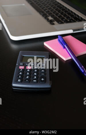 Partial view of a silver keyboard of a laptop with a calculator, and a pink post-it notes with a pencil on a dark wood desk Stock Photo