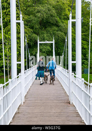 Men walking on River Ness iron footbridge to Ness Island with man wearing killt with dogs Inverness, Scotland, UK Stock Photo