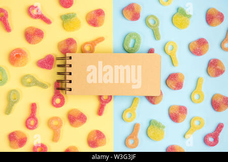 Open blank notebook on background of gummy candies. Multicolored candies on yellow and blue backgrounds. Top view. Stock Photo