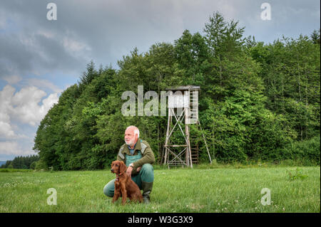 Old hunter sitting in a squatting position in front of his hunting pulpit along with his Irish Setter puppy and looks over his hunting area.. Stock Photo