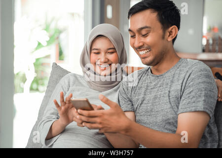 wife pregnant and husband using smartphone together at home while sitting on couch Stock Photo
