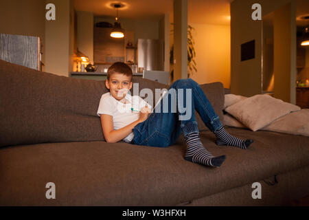 beautiful smiling boy, sitting in couch, looking at camera while writing in his notebook in living room. Stock Photo
