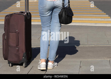 Woman with a suitcase on wheels stands on a pedestrian crossing, rear view. Female legs in blue jeans on the crosswalk, travel, waiting taxi Stock Photo