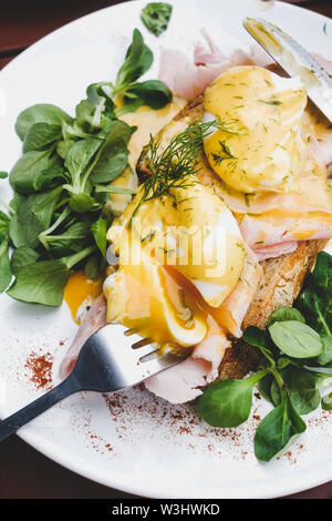 Trendy breakfast set. Flat-lay of poached Eggs Benedict on sourdough bread toast with green salad and pork ham on white plate over cafe table, close-u Stock Photo