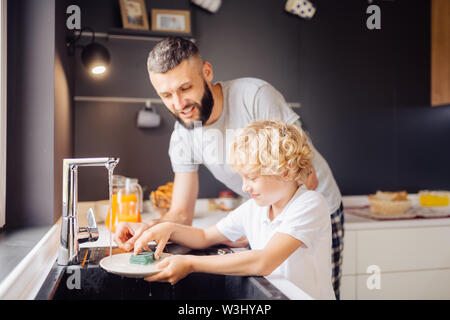 Washing dishes. Joyful nice boy standing near the sink while washing the dishes together with his father Stock Photo