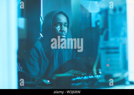 Serious tired black man in hoodie sitting in dark blue room and typing on computer keyboard while working on system software Stock Photo