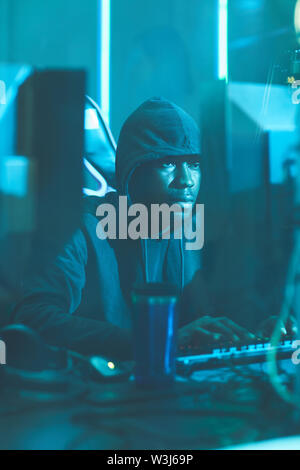 Serious young Afro-American hacker in hoodie sitting at table and typing on computer keyboard while composing data virus code Stock Photo