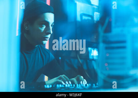 Serious concentrated young Jewish hacker in beanie hat sitting at table in dark room and typing on keyboard while breaking into server data Stock Photo