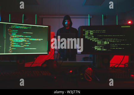 Serious young bearded hacker in hoodie standing in server room with computers and using laptop while getting into computer system Stock Photo