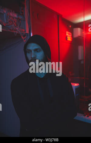 Serious handsome young bearded hacker in hoodie leaving server room with broken Ethernet hub and red light in background Stock Photo