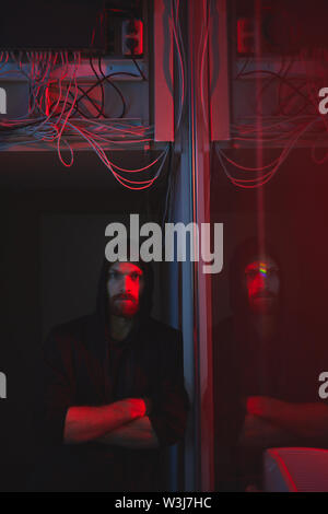Portrait of serious confident young bearded hacker in hoodie leaning on wall in server room with Ethernet hub Stock Photo