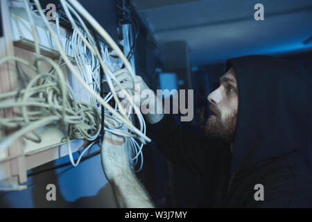 Concentrated young bearded hacker in hoodie checking cables of network server and breaking internet connection while committing cybercrime Stock Photo