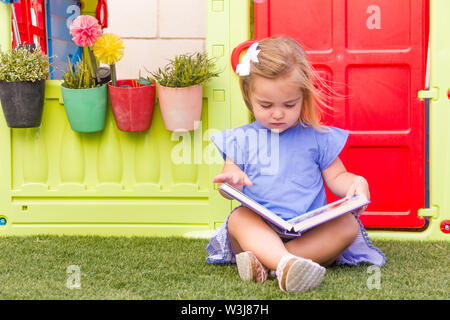 A cute blonde little girl with a blue dress reading a book. She is seated on the grass and there's a colorful toy house on the background. Stock Photo