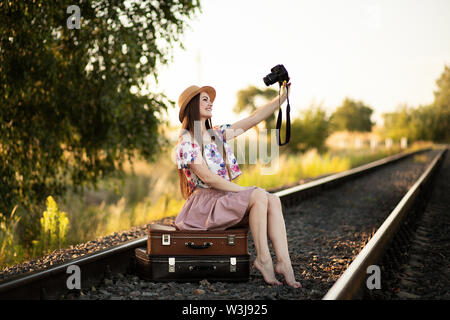 a slender girl in a straw hat is sitting on a suitcases on the railroad tracks, holding a camera and taking pictures of herself. The concept of travel Stock Photo