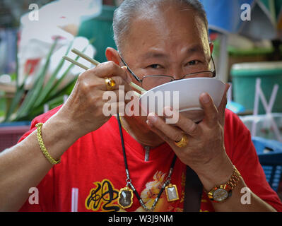 Elderly Thai Chinese man holds a white plastic bowl with noodle soup to his mouth and eats noodles with chop sticks. Stock Photo