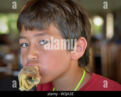 Cute Canadian mixed race boy (Caucasian and Southeast Asian) eats a rolled-up, sweet crêpe (pancake) for breakfast. Stock Photo