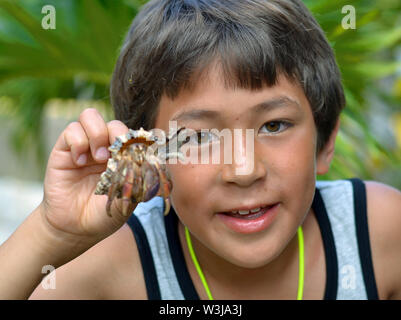 Cute Canadian mixed race boy (Caucasian and Southeast Asian) on vacation in Cuba holds up a hermit crab in a shell. Stock Photo