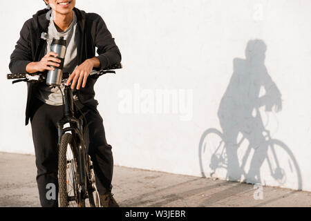 Cropped image smiling asian man riding a bicycle outdoors, drinking water from a bottle Stock Photo