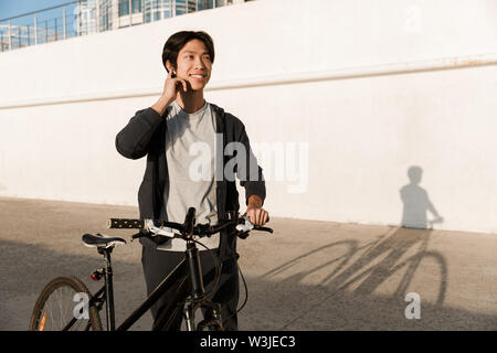 Smiling asian man riding a bicycle outdoors, talking on mobile phone Stock Photo