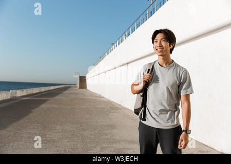 Smiling young asian man wearing backpack spending time outoors at the beach Stock Photo