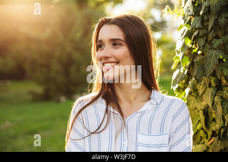 Photo of pretty young woman wearing striped shirt smiling and walking in green park on sunny day Stock Photo