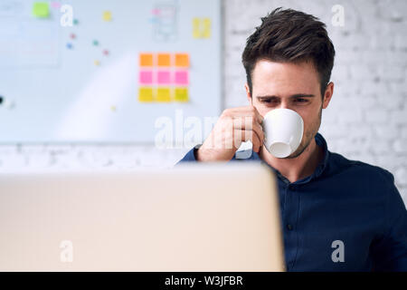 Young man, programmer drinking coffee looking on laptop screen. Stock Photo