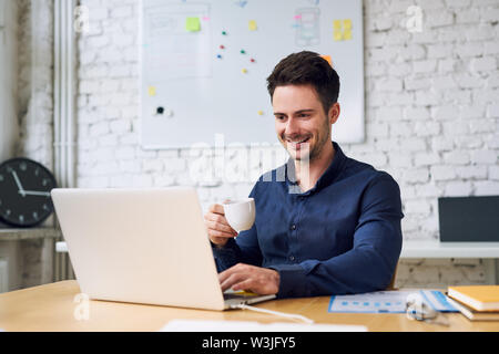 Joyful young startup businessman enjoying coffee and working on laptop in modern home office Stock Photo