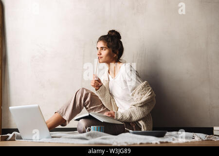 Photo of focused thinking woman wearing leisure clothes writing in diary and using laptop while sitting on floor at home Stock Photo