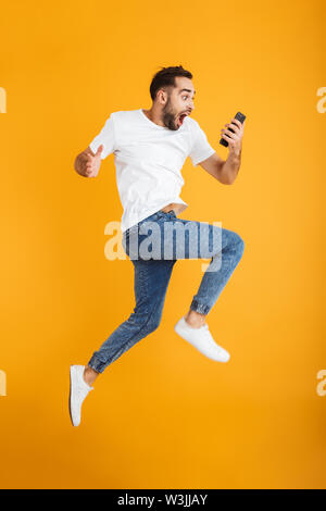 Full length photo of shocked caucasian man having beard wondering and holding cellphone while jumping isolated over yellow background Stock Photo