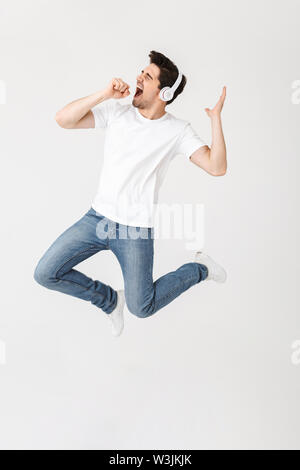 Image of emotional excited young man posing isolated over white wall background listening music with headphones singing jumping. Stock Photo