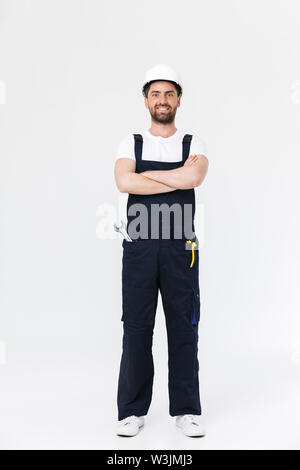 Full length of a confident bearded builder man wearing overalls and hardhat standing isolated over white background, arms folded Stock Photo