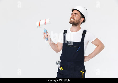 Portrait of a handsome bearded builder man wearing overalls standing isolated over white background, holding paint brush Stock Photo