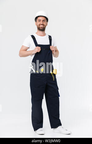 Full length of a confident bearded builder man wearing overalls and hardhat standing isolated over white background, arms folded Stock Photo
