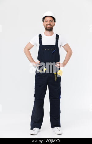 Full length of a confident bearded builder man wearing overalls and hardhat standing isolated over white background, arms folded Stock Photo