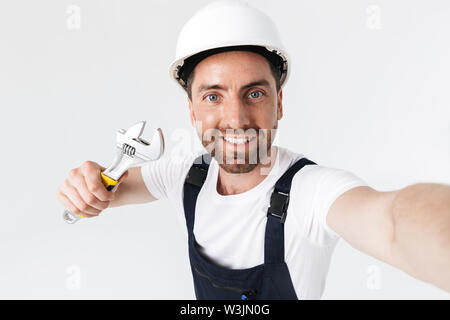 Confident bearded builder man wearing overalls and hardhat standing isolated over white background, showing adjustable wrench, taking a selfie Stock Photo