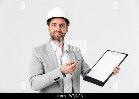 Confident bearded man builder wearing suit and hardhat standing isolated over white background, showing blank notepad Stock Photo