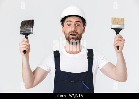 Portrait of a handsome bearded builder man wearing overalls standing isolated over white background, showing two paint brushes Stock Photo
