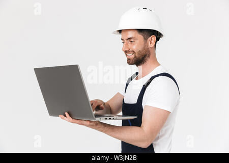 Confident happy bearded builder man wearing overalls and hardhat standing isolated over white background, using laptop computer Stock Photo