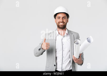 Confident bearded man builder wearing suit and hardhat standing isolated over white background, carrying blueprints, thumbs up Stock Photo