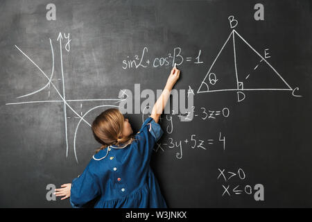 Smart little schoolgirl standing at the blackboard with math graphics, writing on it Stock Photo
